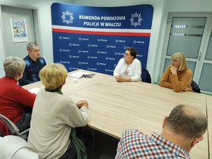 policjant po cywilnemu rozmawia z seniorami, wszyscy siedzą przy stole w tle wyposażenie sali