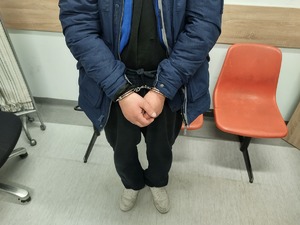 mężczyzna ubrany w kurtkę, na rękach kajdanki w tle ściana i krzesła.