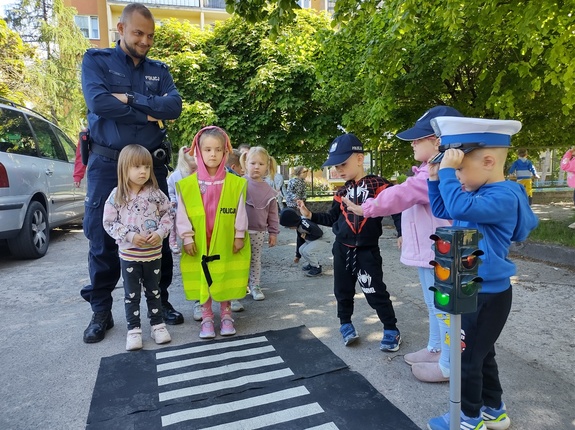 policjant uczy dzieci prawidłowych zachowań na drodze w tle teren przedszkola