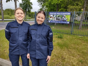 dwie policjantki pozują do zdjęcia w tle baner promujący zawód policjanta wisie na ogrodzeniu