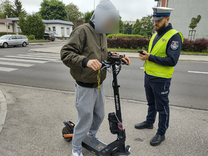 POLICJANT KONTROLUJE OSOBĘ NA HULAJNODZE ELEKTRYCZNEJ W TLE ULICA I POJAZDY