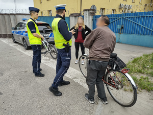POLICJANCI ROZMAWIAJĄ Z ROWERZYSTAMI W TLE OGRODZENIE JEDNOSTKI I ULICA