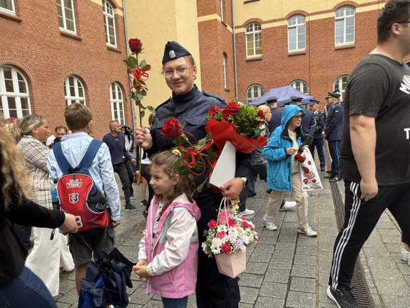 młoda policjantka trzyma w ręce bukiet kwiatów, obok inni ludzie w tle mury komendy policji
