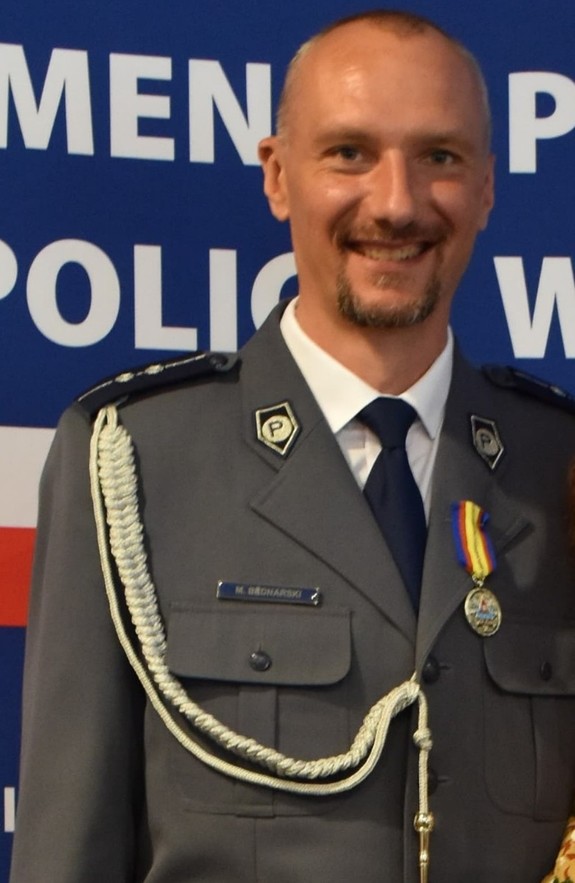 uśmiechnięty policjant na mundurze posiad odznakę w tle baner jednostki