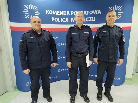 przełożeni wręczają policjantowi awans w tle pomieszczenie i ścianka z logo komendy