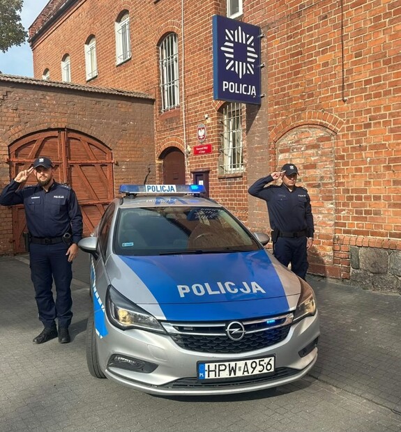 Policjanci stoją przy radiowozie w tle budynek posterunku