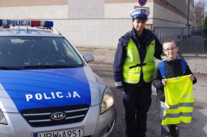 Policjantka wręczyła dziecku kamizelkę odblaskową, w tle radiowóz