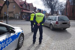 Policjanci stoją na drodze, prowadzą kontrole pojazdów, w tle dwa ososbowe auta i zabudowania