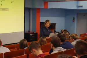 policjantka ma pochyloną głowę, czyta z kartki, w tle ściana sali, przed policjantką siedzący uczniowie
