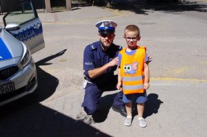 policjant klęczy obok chłopca w kamizelce odblaskowej,  w tle zabudowania