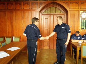komendant ściska rękę policjantowi z gratulacjami, w tle ściana drweniana i drzwi od sali
