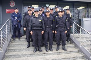 policjanci stoją na schodach przed wejściem głównym do komendy, w tle tablica informacyjna jednostki