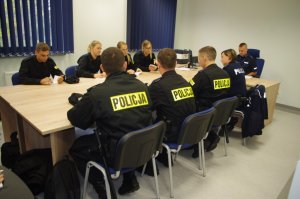 policjanci w sali odpraw, siedzą za stołem,  w tle rolety na oknach