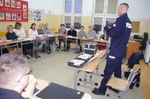 policjant prezentuje materiały edukacyjne, w tle nauczyciele