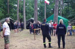 na pierwszym planie policjantka rozmawia z wypoczywającymi, w tle obóz w lesie