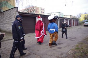 Mikołaj i policyjna maskotka podchodzą do Pani kierownik DPS, w tle garaże i zaparkowany samochód