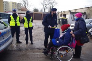 policjant wręcza odblask kobiecie na wózku inwalidzkim, w tle policjanci i zabudowania