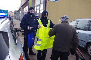 policjantka zakłada mężczyźnie z rowerem odblaskową kamizelkę, w tle zabudowania