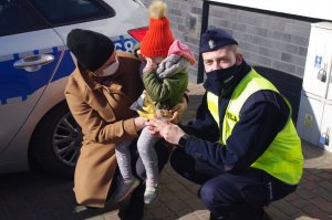 policjant wręcza maskotkę dziewczynce, obok mama dziecka, w tle radiowóz